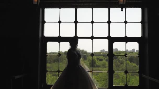 ウェディングドレスを着た花嫁が大きな窓で回転している 窓から光が部屋に入ってくる 花嫁の手に花束 — ストック動画