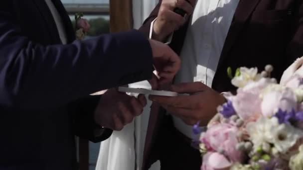 新郎は結婚式で花嫁に結婚指輪を置きます 白いチュールのアーチを背景に 川が流れ 結婚式の花束の手に — ストック動画