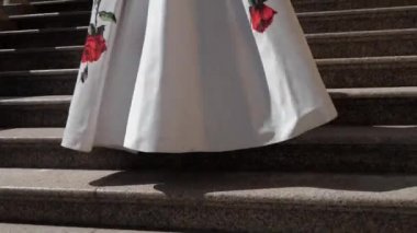 Bir kadın kırmızı çiçeklerle süslenmiş beyaz bir etek içinde merdivenlerden yukarı yürüyor. Taştan yapılmış basamaklar ve korkuluklar. Güneşli gün, güller.