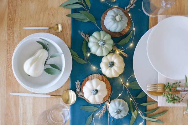 Blue, teal and cream pumpkins Thanksgiving table decorations with white tableware, brass kitchenware, dry eucalyptus leaves, greenery and string lights