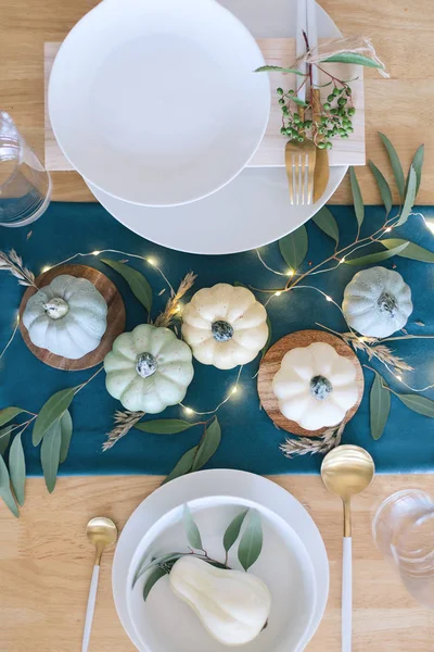 Blue, teal and cream pumpkins Thanksgiving table decorations with white tableware, brass kitchenware, dry eucalyptus leaves, greenery and string lights