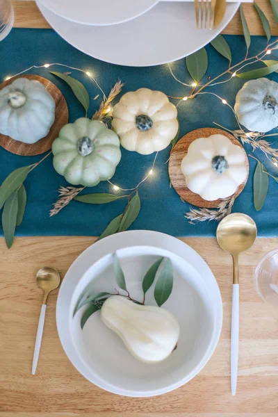 Blue, teal and cream pumpkins Thanksgiving table decorations with white tableware, brass kitchenware, dry eucalyptus leaves, greenery and string lights