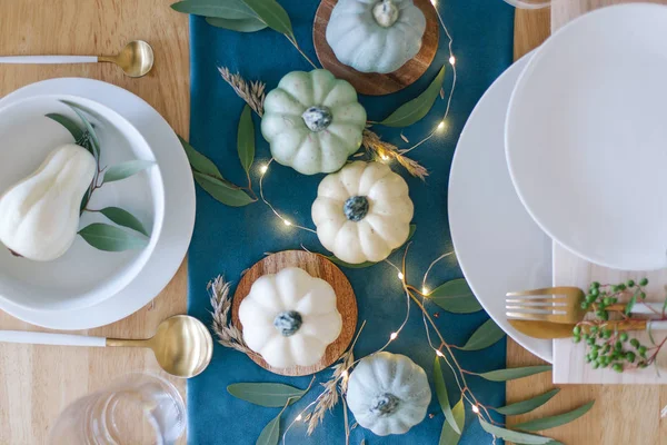 Blue, teal and cream pumpkins Thanksgiving table decorations with white tableware, brass kitchenware, dry eucalyptus leaves, greenery and string lights