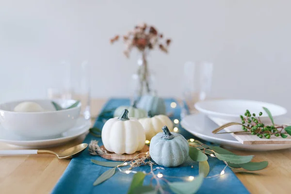 Blue, teal and cream pumpkins Thanksgiving table decorations with white tableware, brass kitchenware, dry eucalyptus leaves, greenery and string lights