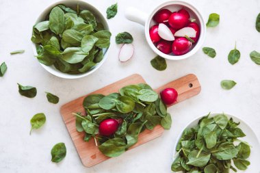 Bebek ıspanak yaprakları ve beyaz kaselerde turplar ve beyaz mutfak masasında tahta kesme. Yaz ve bahar taze sebze salata tarifi.