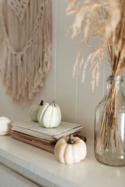 Thanksgiving home  decor - white wooden fireplace with wall macrame, cream and beige pumpkins, pine cones garland and rustic dry wheet bouquet. House fall decoration.