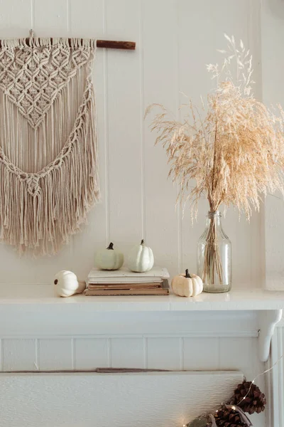 Thanksgiving home  decor - white wooden fireplace with wall macrame, cream and beige pumpkins, pine cones garland and rustic dry wheet bouquet. House fall decoration.