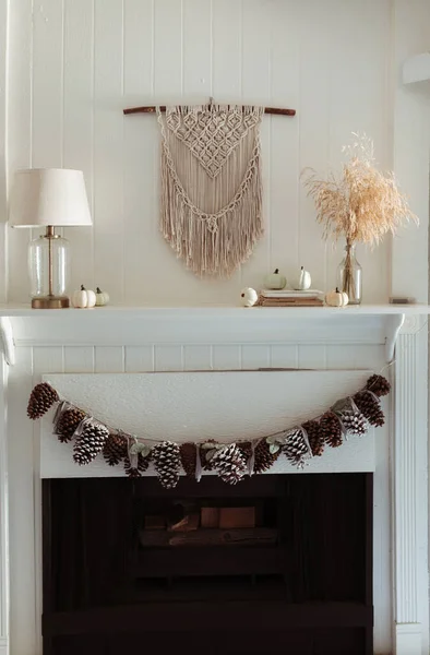 Thanksgiving home  decor - white wooden fireplace with wall macrame, cream and beige pumpkins, pine cones garland and rustic dry wheet bouquet. House fall decoration.