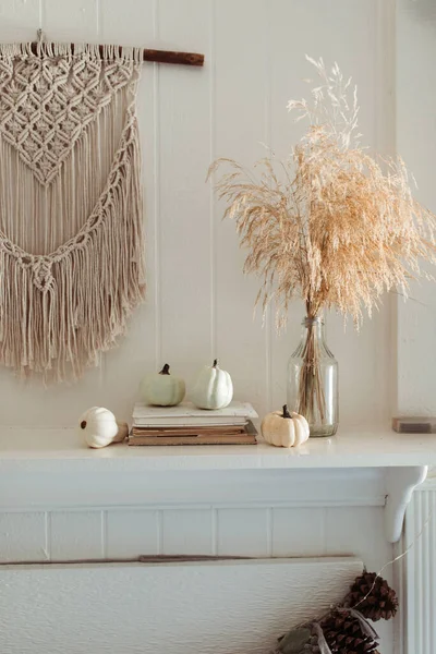 Thanksgiving home  decor - white wooden fireplace with wall macrame, cream and beige pumpkins, pine cones garland and rustic dry wheet bouquet. House fall decoration.