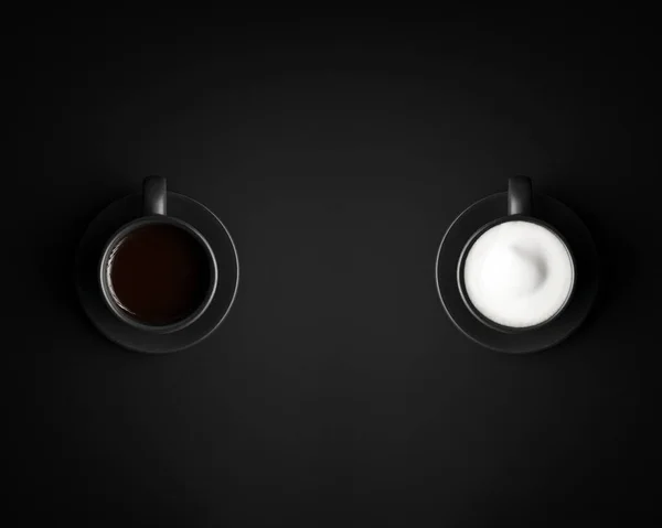 Duas xícaras pretas e pires. Um com cappuccino, o outro com café preto . — Fotografia de Stock