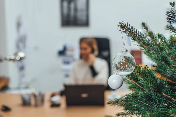 Ofiste süslenmiş Noel ağacı, bulanık arka planda iş kadını dizüstü bilgisayarla çalışıyor.