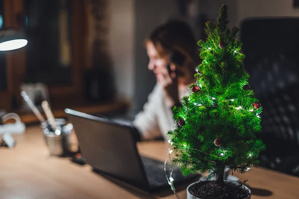 Ofiste süslenmiş Noel ağacı, bulanık arka planda iş kadını dizüstü bilgisayarla çalışıyor.