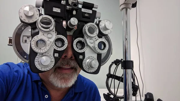 Mavi tişörtlü keçi sakallı adam diopter makinesiyle göz muayenesi yaptırıyor.