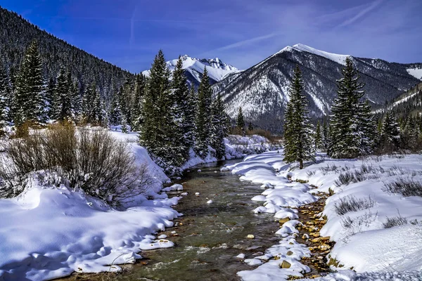 Snow covered peaks in the distance covered with freshly fallen snow in mountain valley with river flowing through alpine forest with trees and rocks