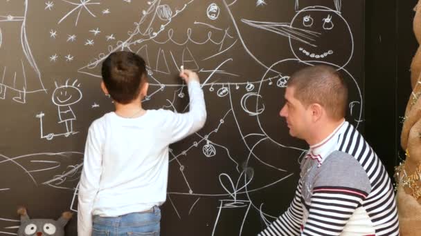 Αγόρι με πατέρα χρωματίζει μια εικόνα στον τοίχο με κιμωλία σε ένα θέμα Χριστουγέννων — Αρχείο Βίντεο