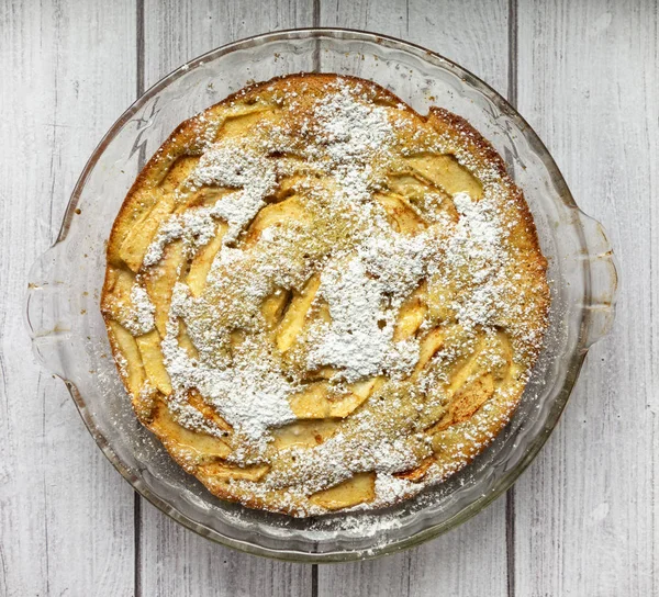 A homemade apple cake in a glass pie pan dusted with confectioner sugar on a light wood background.