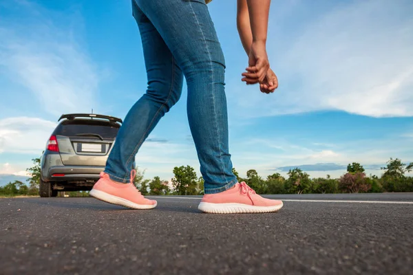 Women wearing jeans binding sneakers on the highway, car parked along the way in the daytime, the bright blue sky and clouds background