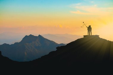 Gün batımında bir dağın tepesinde selfie çeken bir aşığın silüeti.