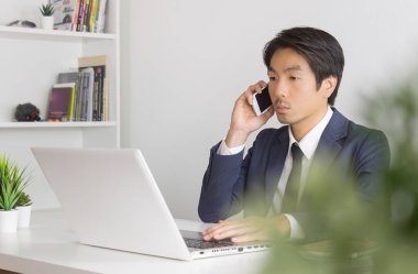 Resmi Takım Elbiseli Asyalı İşadamı Akıllı Telefonu ve Ofiste Dizüstü Bilgisayarı Kullanıyor