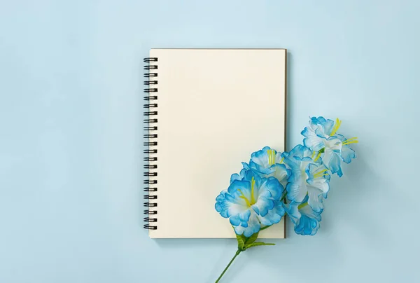 Spiral Notebook or Spring Notebook in Unlined Type and Blue Flowers at Bottom Right on Blue Pastel Minimalist Background. Spiral Notebook Mockup on Center Frame