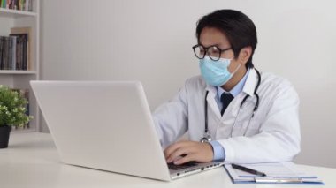 Genç Asyalı doktor önlüklü ya da gecelikli steteskop gözlüklü ve maske takan ofiste dizüstü bilgisayar kullanan.