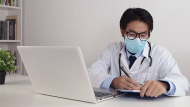 Laboratuar önlüklü genç Asyalı doktor ya da steteskoplu cüppe Gözlük ve Yüz Maskesi Muayenesi Hastası Ofiste Video Çağrısı ya da Video Konferansı
