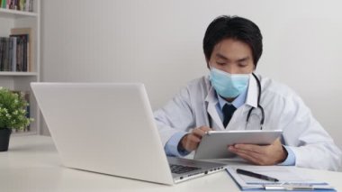 Laboratuvar önlüklü genç Asyalı doktor ya da gecelikli stetoskop Yüzü Maske Giyen Online Toplantı ya da Tablet 'in Video Konferansı ve Ofiste Laptop