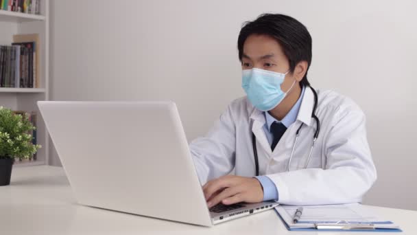 Lab Coatの若いアジアの医師の男性またはオフィスの医師テーブルのラップトップコンピュータを使用して聴診器摩耗顔マスクでガウン — ストック動画