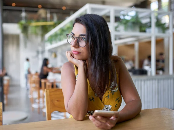 Candid lifestyle portrait of beautiful hispanic millennial woman looking out window holding cellphone in modern trendy and bright coffee shop with plants — Stock Photo, Image