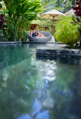 Bali tropikal otel ve spa lüks havuz yakınında modern sandalyede uzanmış genç kafkas çift samimi çekim