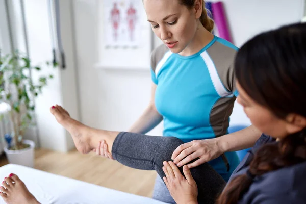 Blanke vrouw fysiotherapeut strectching het been en de knie van een mid-Adult Chinese vrouwelijke patiënt zittend op een massagetafel — Stockfoto