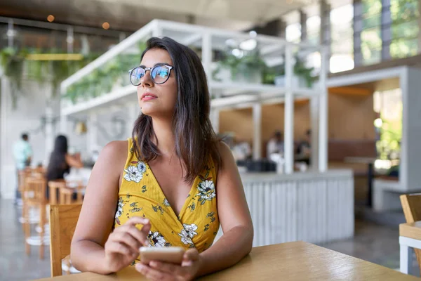 Ehrliches Lifestyleporträt der schönen hispanischen Millennial-Frau, die mit dem Handy in einem modernen trendigen und hellen Café mit Pflanzen arbeitet — Stockfoto
