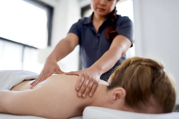 Chińska kobieta masażystka daje leczenie atrakcyjny blond klienta na stole do masażu w jasnym biurze medycznym — Zdjęcie stockowe