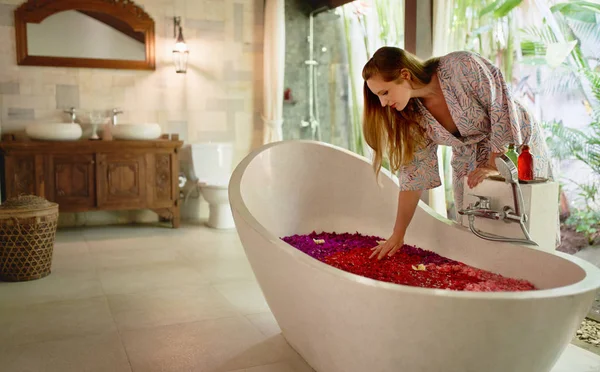 Pretty natural female touching flower petals floating in luxurious bathtub of tropical spa and wellness centre — Stock Photo, Image