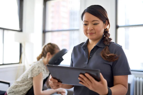 portrait of a Chinese woman massage therapist holding a tablet while filling the file of an attractive blond client at her workplace in a bright office