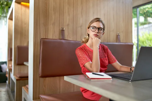 Primo piano stile di vita ritratto di pensoso bionda caucasico millennial vlogger nel suo 20s indossare occhiali lavorando su computer portatile in un ristorante moderno — Foto Stock
