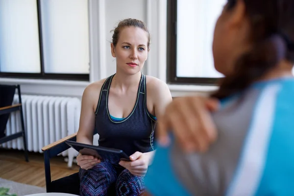 Mulher branca fisioterapeuta conversando com uma paciente chinesa média adulta e tomando notas em um comprimido durante uma consulta de avaliação de fitness — Fotografia de Stock