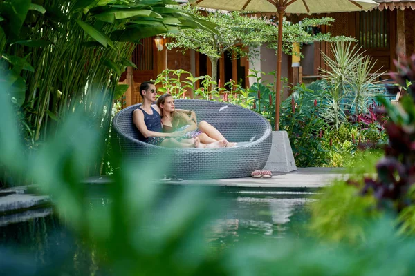 Candid shot fiatal kaukázusi pár heverészett a modern szék közelében luxus medence trópusi szálloda és gyógyfürdő Balin — Stock Fotó