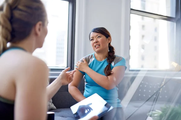 Mulher branca fisioterapeuta conversando com uma paciente chinesa média adulta e tomando notas em um comprimido durante uma consulta de avaliação de fitness — Fotografia de Stock