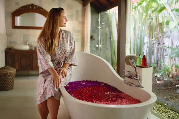 Beautiful millennial woman relaxing outside large bathtub filled with flower petals in tropical spa setting — Stock Photo, Image