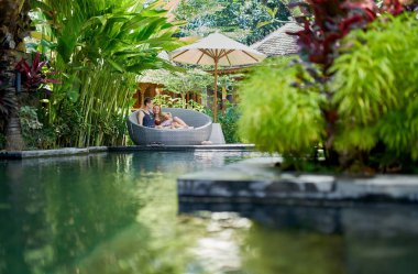 Bali tropikal otel ve spa lüks havuz yakınında modern sandalyede uzanmış genç kafkas çift samimi çekim