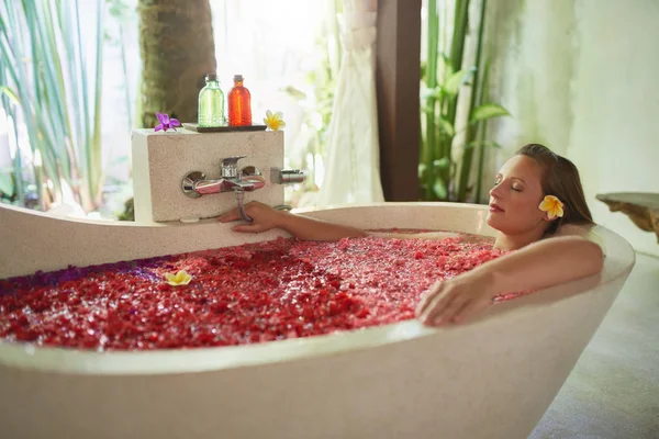 Natural millennial woman being pampered in luxurious white bathtub filled with flower petals in tropical resort and wellness centre — Stock Photo, Image