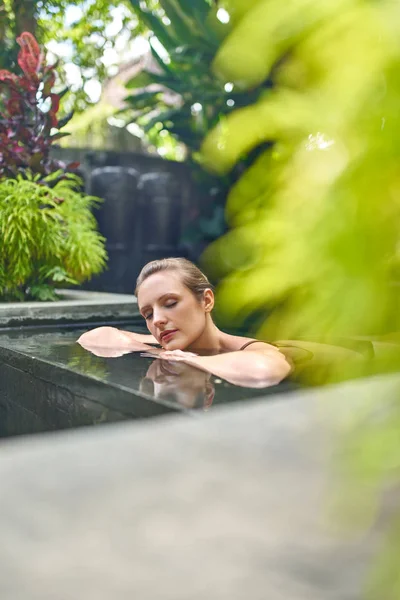 Healthy woman relaxing outdoors in sunny and plant surrounded swimming pool of luxurious hotel during tropical vacation in Bali — Stock Photo, Image