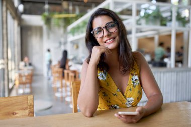 Modern restoranda oturmuş kameraya gülümseyen, kendine güvenen, güzel, doğal esmer İspanyol bir vlogger 'ın hayat tarzı portresi.
