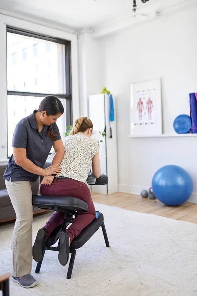 Chinesische Masseurin gibt einer attraktiven blonden Kundin an ihrem Arbeitsplatz in einem hellen Büro eine Nacken- und Rückendruckbehandlung — Stockfoto