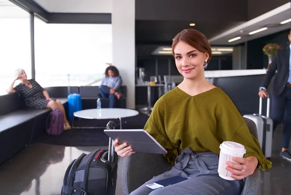 Millennial female traveller with coffee sitting in modern airport terminal holding tablet looking at camera — ストック写真
