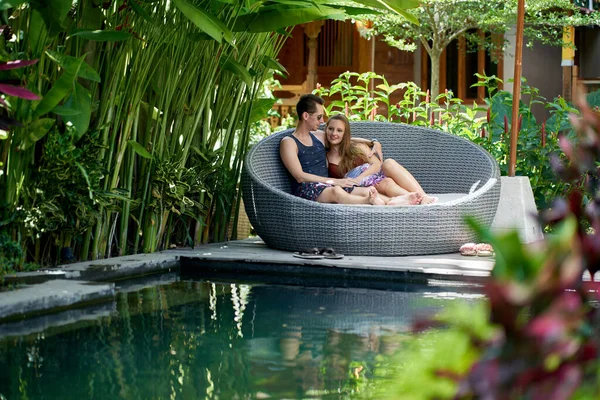 발리의 열대 호텔과 스파의 고급스러운 수영장 근처 현대 의자에서 느긋하게 휴식을 즐기는 젊은 백인 부부의 솔직한 샷 — 스톡 사진