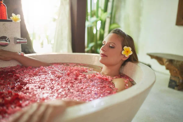 Beautiful millennial woman with tropical flower accessory sitting in large bathtub filled with flower petals in luxurious spa setting — Stock Photo, Image