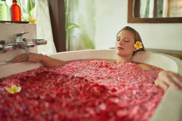 Menina milenar lindo mimando-se na banheira moderna cheia de pétalas de flores em resort tropical e spa — Fotografia de Stock