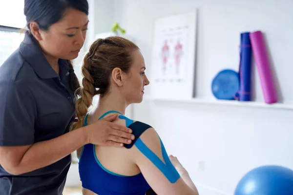 Terapeuta de massagem chinesa aplicando fita kinesio para os ombros e pescoço de um cliente loiro atraente em um escritório médico brilhante — Fotografia de Stock